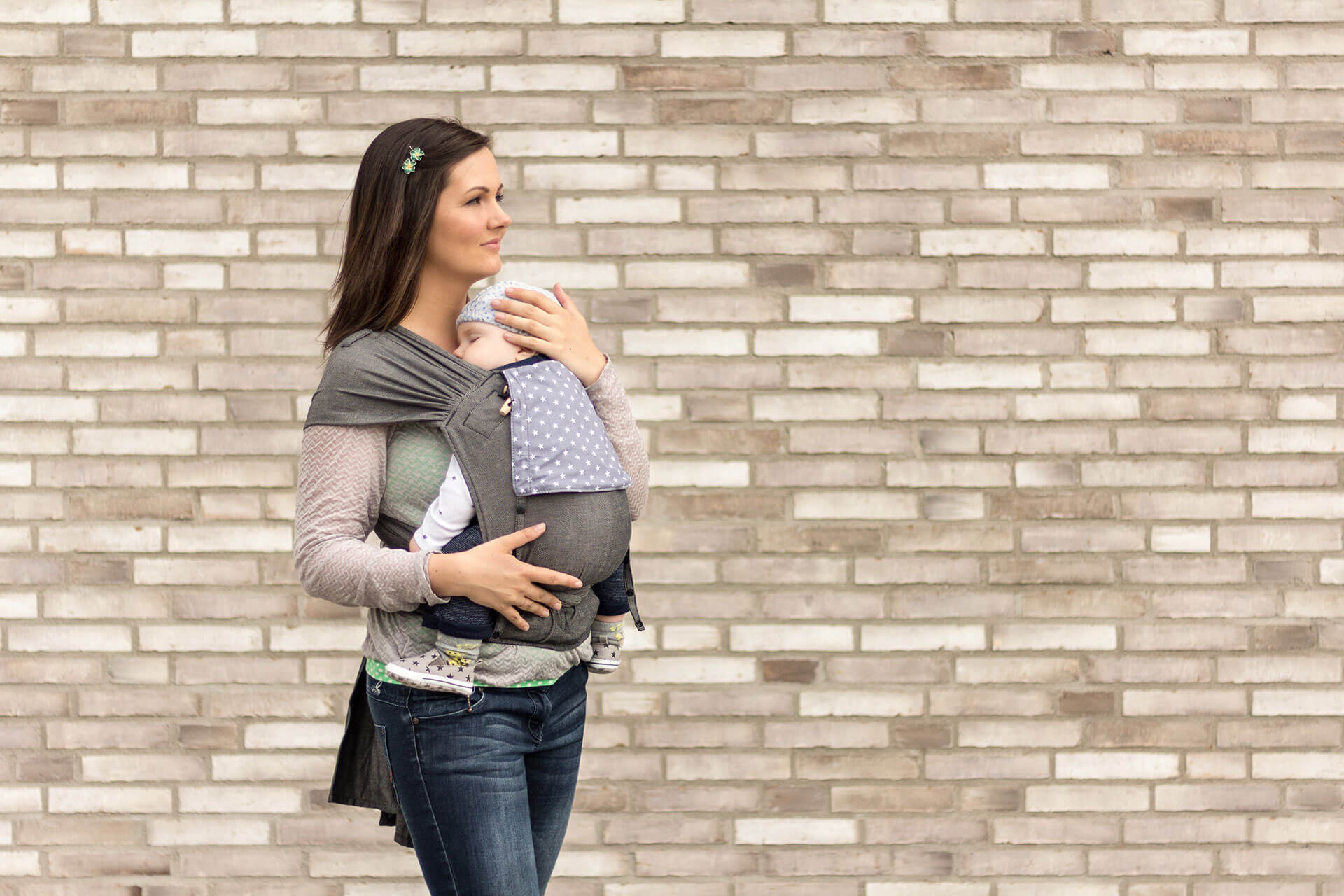 Babytrage-WrapCon Eine Frau trägt ihr Baby in einer WrapCon BAbytrage. Sie steht vor einer BAcksteinmauer. Die Träger der Babytrage sind breit über ihren Schultern aufgefächert für eine bessere Gewichtsverteilung.
