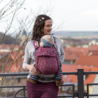 Eine Frau mit dunklen Haaren trägt ihr Baby vor dem Bauch in einem weinrot gemusterten Fräulein Hübsch Mei Tai. Im Hintergrund sieht man Himmel und Häuser.