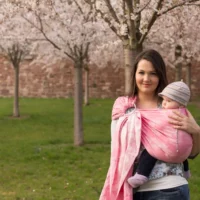 A woman carries her baby in a pink ring sling on her hip. She stands on a green meadow in front of flowering trees.
