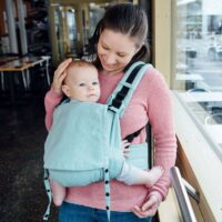 Eine Mutter trägt ihr Baby vor dem Bauch in einer Fräulein Hübsch Fullbuckle Babytrage der Größe Onesize. Die Trage ist helltürkis, Baby und Mutter stehen vor einem FEnster und schauen in die Kamera. Die Mutter lächelt.