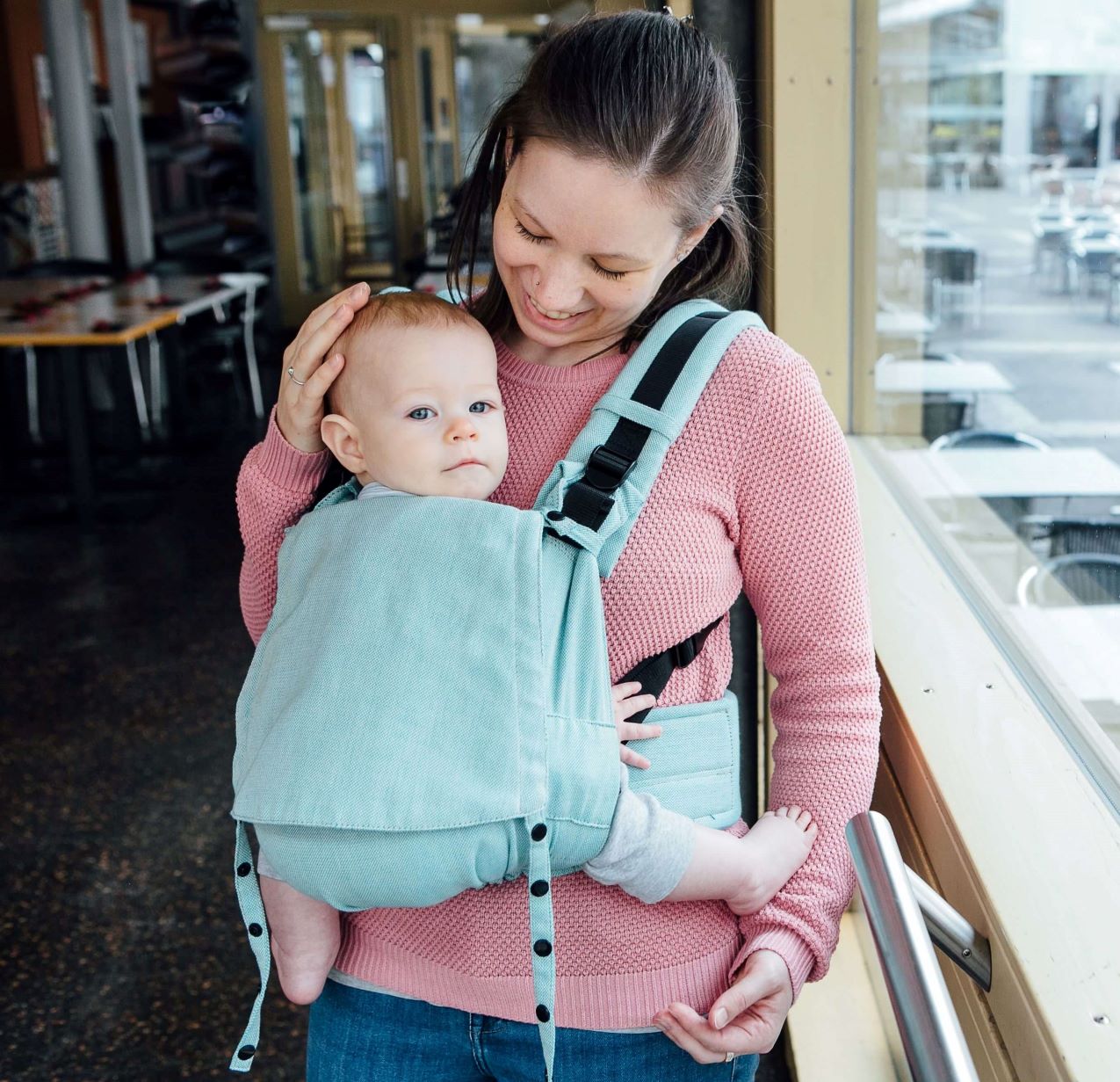 Fullbuckle Babytrage_die richtige Babytrage kaufen Eine Mutter trägt ihr Baby vor dem Bauch in einer Fräulein Hübsch Fullbuckle Babytrage der Größe Onesize. Die Trage ist helltürkis, Baby und Mutter stehen vor einem FEnster und schauen in die Kamera. Die Mutter lächelt.