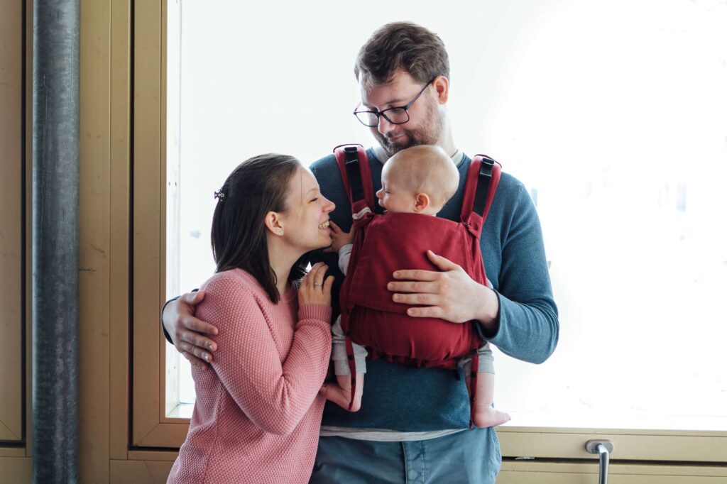 Ein Vater trägt sein Kind in einer Fräulein Hübsch Fullbuckle Babytrage vor dem Bauch, die Mutter steht daneben und lächelt das Baby an