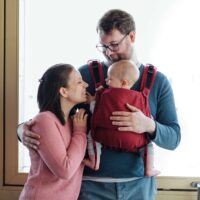 Ein Vater trägt sein Kind in einer Fräulein Hübsch Fullbuckle Babytrage vor dem Bauch, die Mutter steht daneben und lächelt das Baby an