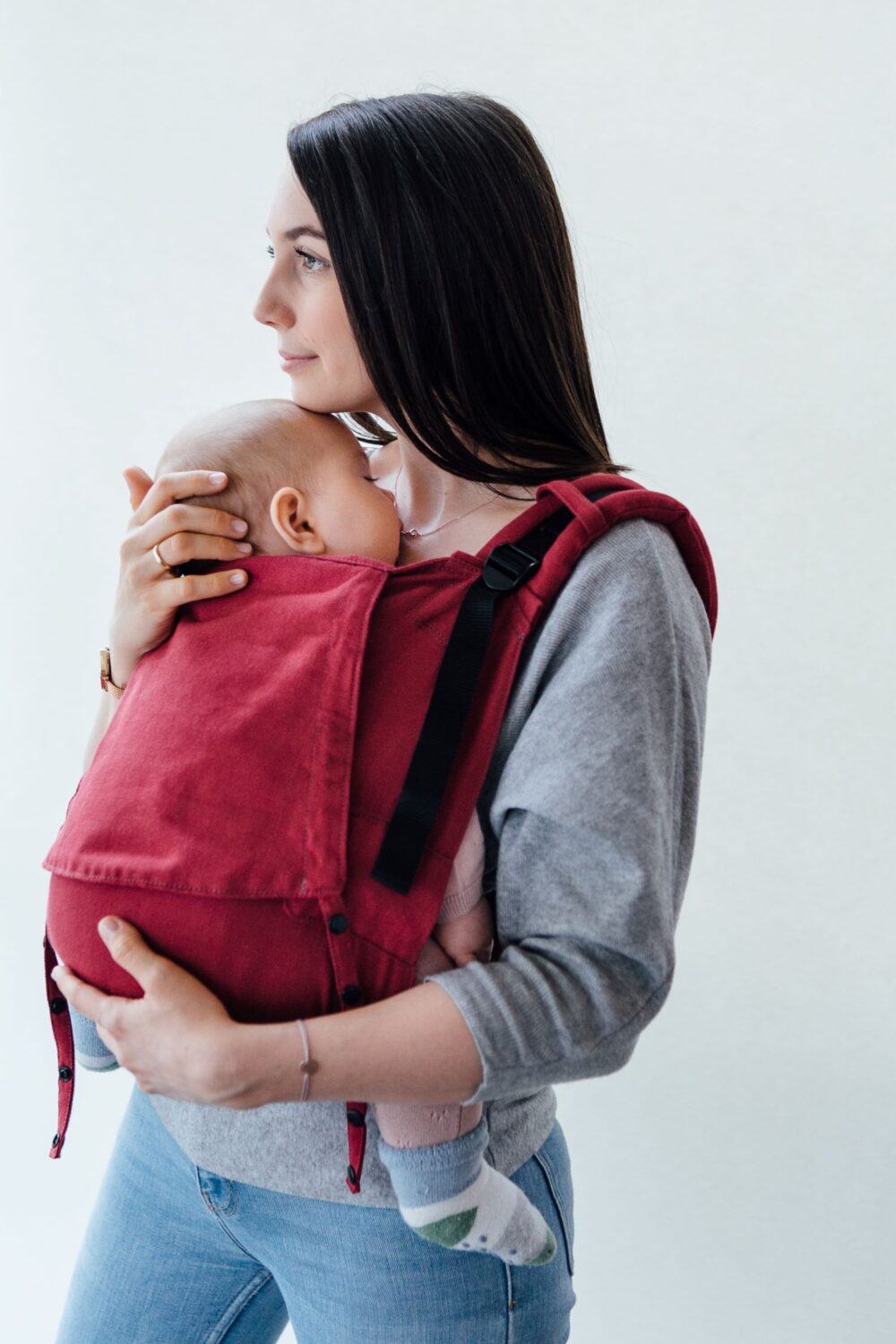 _kalinkaphoto_FrlHübsch-9_komp Eine Mutter trägt ihr Baby vor dem Bauch in einer roten Fräulein Hübsch Fullbuckle Babytrage. Sie steht vor einer weißen Wand und sieht nach links. Das Baby schläft in der Babytrage.