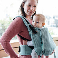 Eine Mutter trägt ihr Baby vor dem Bauch in einer Fräulein Hübsch Fullbuckle Babytrage der Größe Onesize. Die Trage ist helltürkis, Baby und Mutter stehen vor einem FEnster und schauen in die Kamera. Die Mutter lächelt.