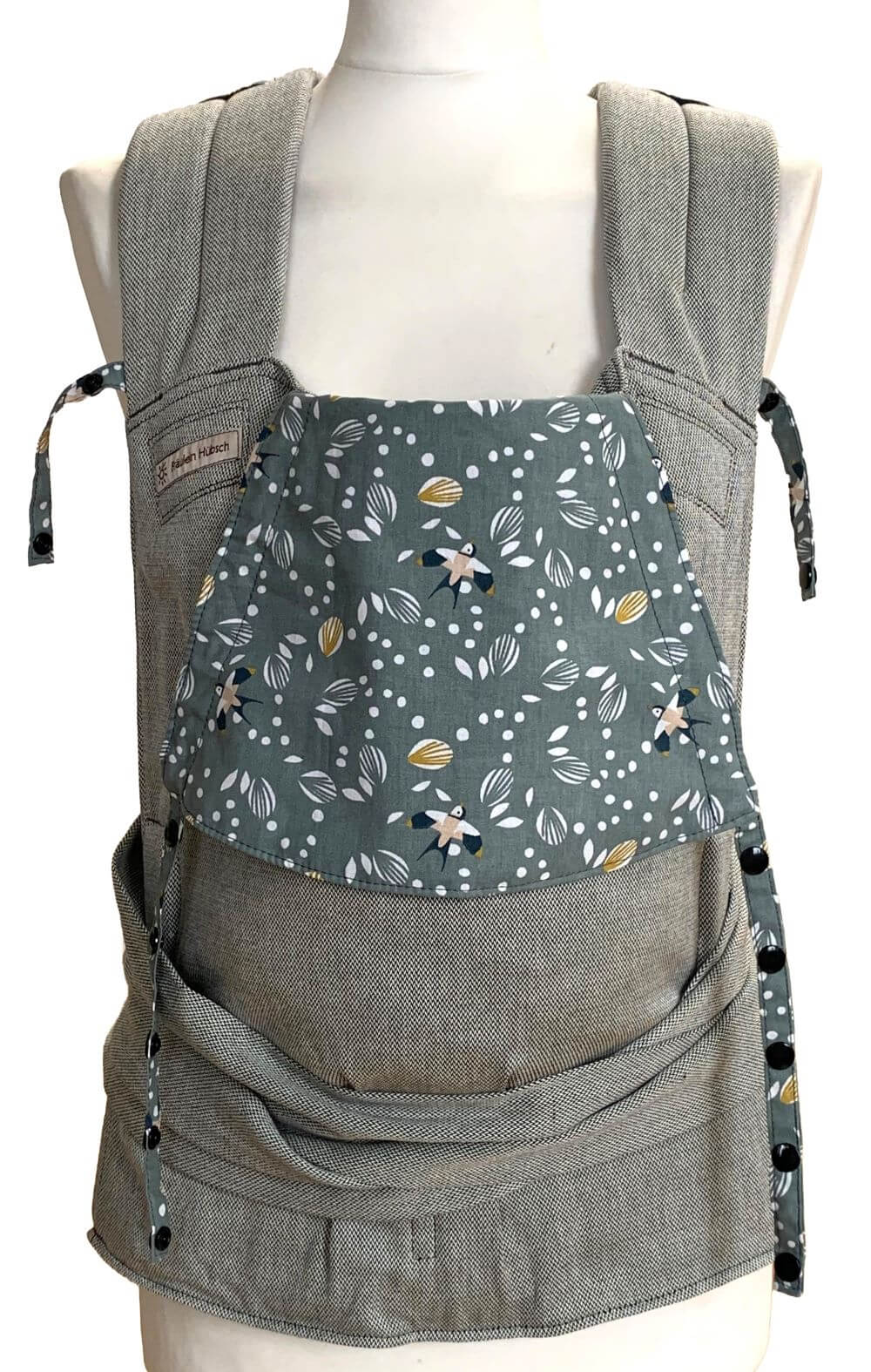 Babytrage in Hellgrau mit Kopfstütze mit salbeigrünem Hintergrund, auf dem weiße Blätter und Schwalben in Weiß, Schwarz und Ocker zu sehen sind.