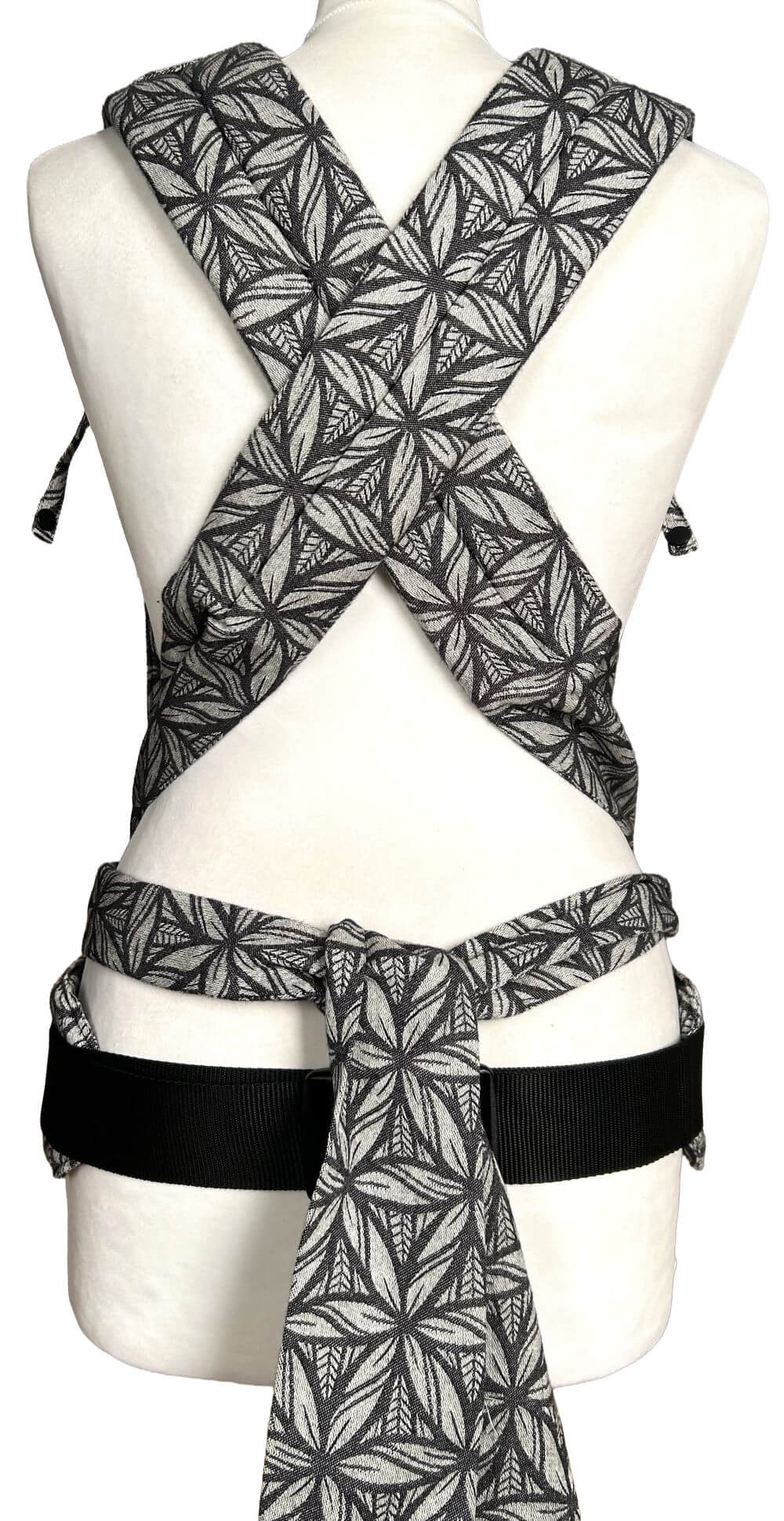 Babytrage mit Schulterträgern zum Binden und Hüftgurt mit Schnalle in grafischem Muster aus Weiß und Schwarz auf einer Schneiderbüste. Ansicht von hinten, man sieht die gebundenen Träger.