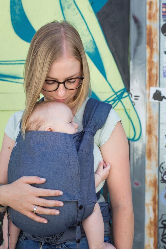 Eine Frau trägt ein Baby in einer Fräulein Hübsch SoftTai Babytrage. Das Baby sitzt vor dem Bauch, die Frau steht vor einer bunten Graffiti Wand und küsst ihr Baby auf den Kopf.