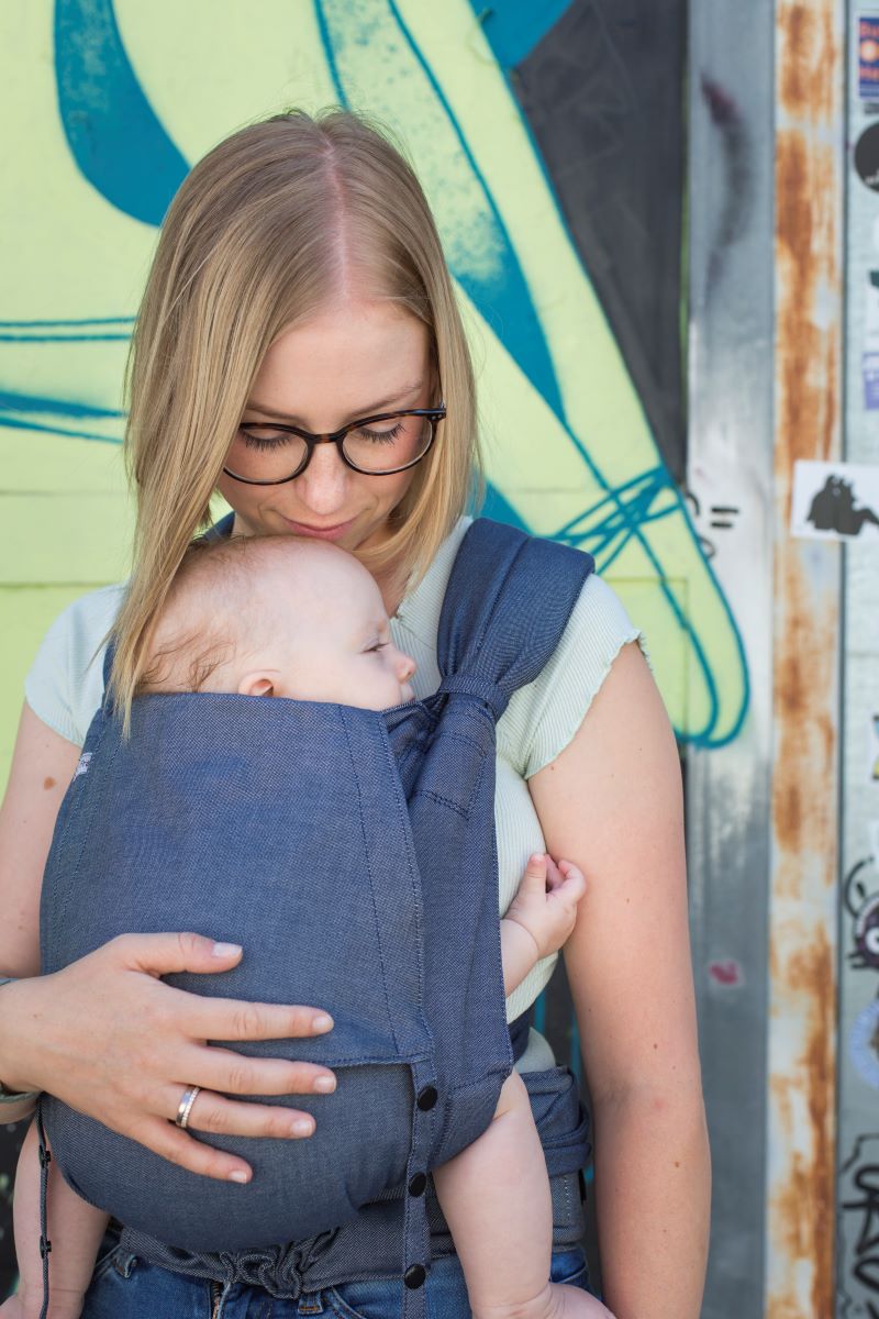 Fräulein Hübsch Babytrage SoftTai BAbysize Denim Eine Frau trägt ein Baby in einer Fräulein Hübsch SoftTai Babytrage. Das Baby sitzt vor dem Bauch, die Frau steht vor einer bunten Graffiti Wand und küsst ihr Baby auf den Kopf.