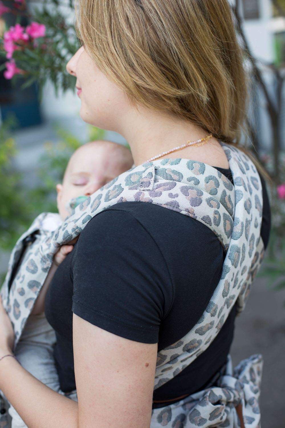 Blonde Frau mit schwarzem T-Shirt trägt ihr BAby in einer Babytrage mit Leoprint. Sie ist von schräg hinten zu sehen und man sieht, wie die Träger der Babytrage auf dem Rücken überkreuzt sind für mehr Tragekomfort.