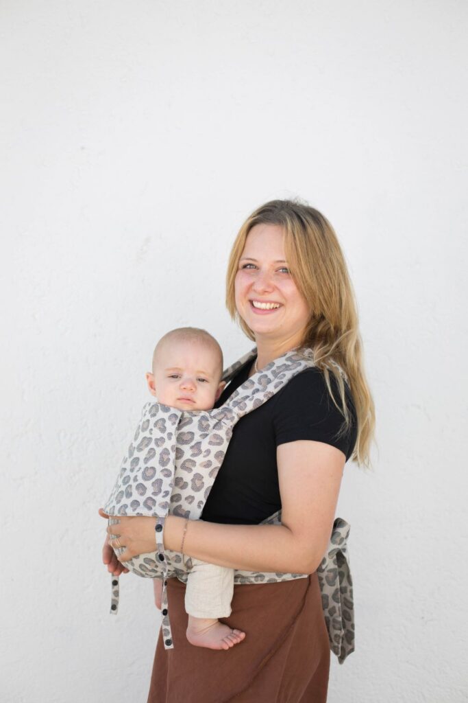 Blonde Frau mit schwarzem T-Shirt trägt ihr Baby sicher in einer Babytrage mit Leoprint und lächelt in die Kamera. Sie achtet auf freie Atemwege und auf die Sicherheit ihres BAbys in der Babytrage.