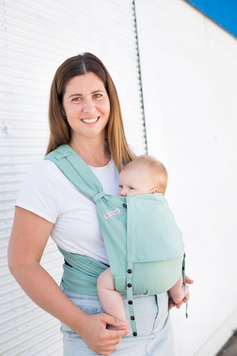 Fräulein Hübsch Babytrage SoftTai BAbysize Jade Eine Frau trägt ein Baby in einer Fräulein Hübsch SoftTai Babytrage. Das Baby sitzt vor dem Bauch, die Frau steht vor einer weißen Wand und sieht nach links.