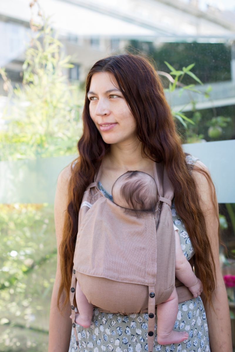 Fräulein Hübsch Babytrage SoftTai BAbysize Toffee Eine Frau trägt ein Baby in einer Fräulein Hübsch SoftTai Babytrage. Das Baby sitzt vor dem Bauch, die Frau steht vor einer Glastür neben Pflanzen und sieht nach links.