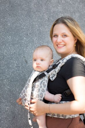 Eine blonde Frau trägt ihr Baby vor dem Bauch in einer Fullbuckle Onesize Babytrage von Fräulein Hübsch im Muster Leoprint. Mutter und Baby stehen vor einer grauen WAnd und lächeln in die Kamera.