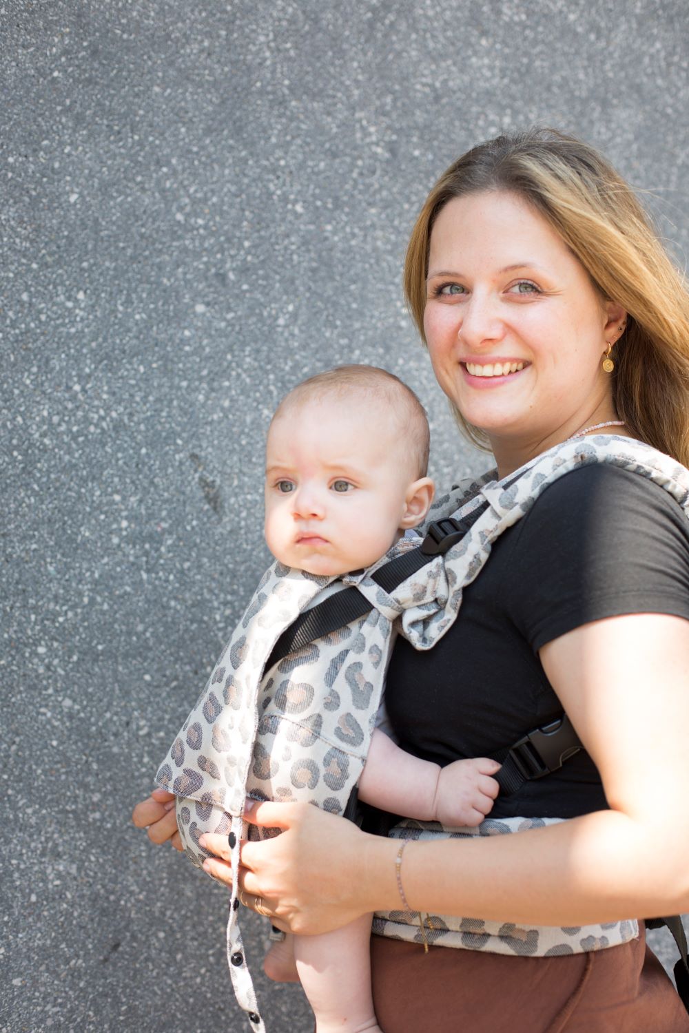 Fräulein Hübsch Fullbuckle Onesize Leoprint Eine blonde Frau trägt ihr Baby vor dem Bauch in einer Fullbuckle Onesize Babytrage von Fräulein Hübsch im Muster Leoprint. Mutter und Baby stehen vor einer grauen WAnd und lächeln in die Kamera.