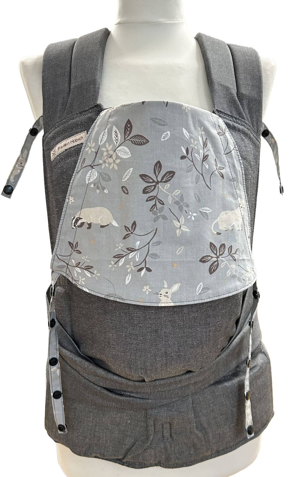 Babytrage von Fräulein Hübsch in Dunkelgrau mit mittelgrauer Kopfstütze mit kleinen Tieren wie Hermelin, Biber und Hase und Zweigen mit Blättern in Beige und Dunkelgrau. Die Babytrage schließt am Bauchgurt mit Schnalle und die SChulterträger sind zum Binden.