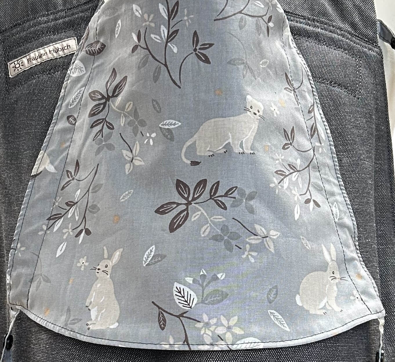 Detail des Kopfstützen-Stoffes: mittelgrauer Stoff mit dunkelgrauen und beigen Zweigen mit Blättern zwischen denen ein Dachs, ein Hermelin und ein Hase zu sehen sind.