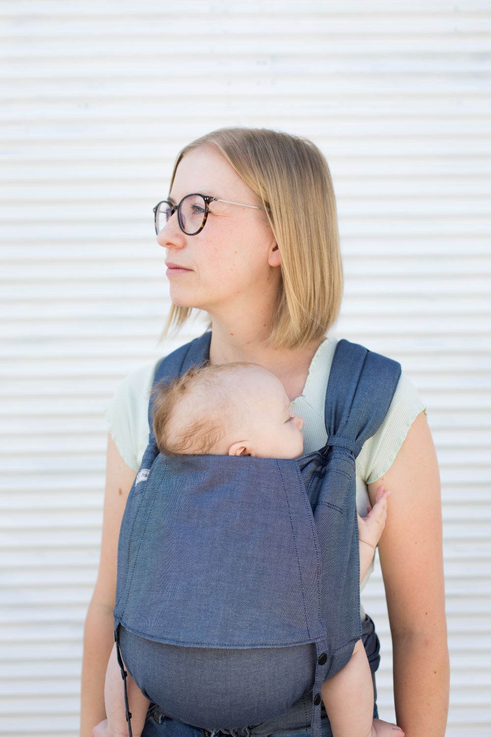_69A8709_komp Eine Frau trägt ein Baby in einer Fräulein Hübsch SoftTai Babytrage. Das Baby sitzt vor dem Bauch, die Frau steht vor einer weißen Wand und sieht nach links.
