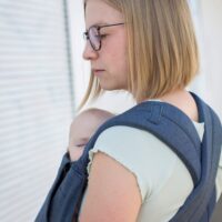 Eine blonde Frau trägt ihr Baby in einer Babytrage SoftTai von Fräulein Hübsch. Man sieht sie von schräg hinten, die Träger sind am Rücken überkreuzt.