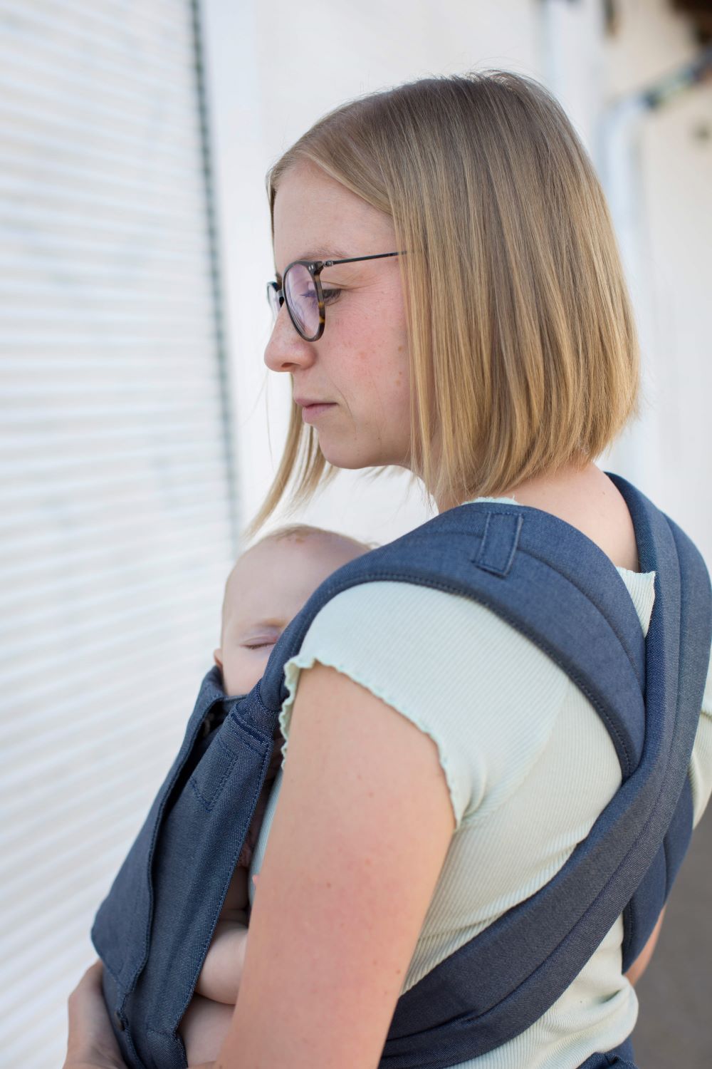_69A8730_komp Eine blonde Frau trägt ihr Baby in einer Babytrage SoftTai von Fräulein Hübsch. Man sieht sie von schräg hinten, die Träger sind am Rücken überkreuzt.
