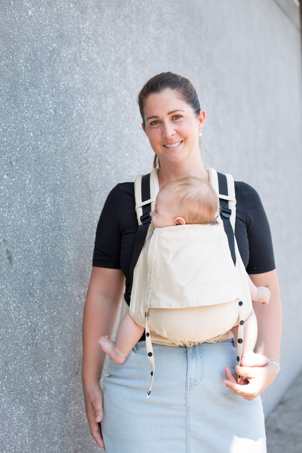 Fräulein Hübsch Babytrage Fullbuckle Newborn Farbe Sand 1 Eine Mutter trägt ihr Baby in der Babytrage Fullbuckle Newborn vor dem Bauch. Sie steht vor einer grauen Wand und lächelt in die Kamera. Das Bild ist frontal aufgenommen.