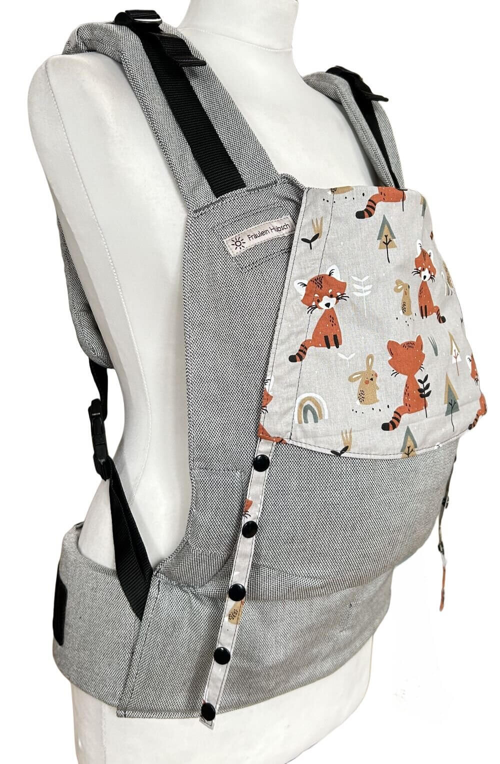 Fullbuckle Newborn Foxes seitlich Fullbuckle Rucksacktrage Größe Newborn in Hellgrau mit einer Kopfstütze mit Füchsen auf Grau einer Schneiderbüste. Alle Gurte schließen mit Schnallen wie bei einem Rucksack, kein Binden.