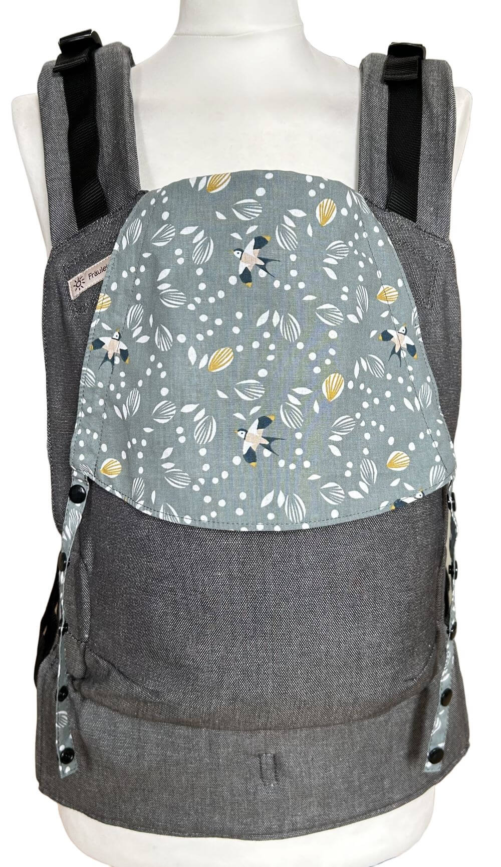 Fullbuckle Newborn Swallows Frontal Fullbuckle Rucksacktrage Größe Newborn in Dunkelgrau mit einer Kopfstütze mit Schwalbenmuster auf Salbeigrün auf einer Schneiderbüste. Alle Gurte schließen mit Schnallen wie bei einem Rucksack, kein Binden.