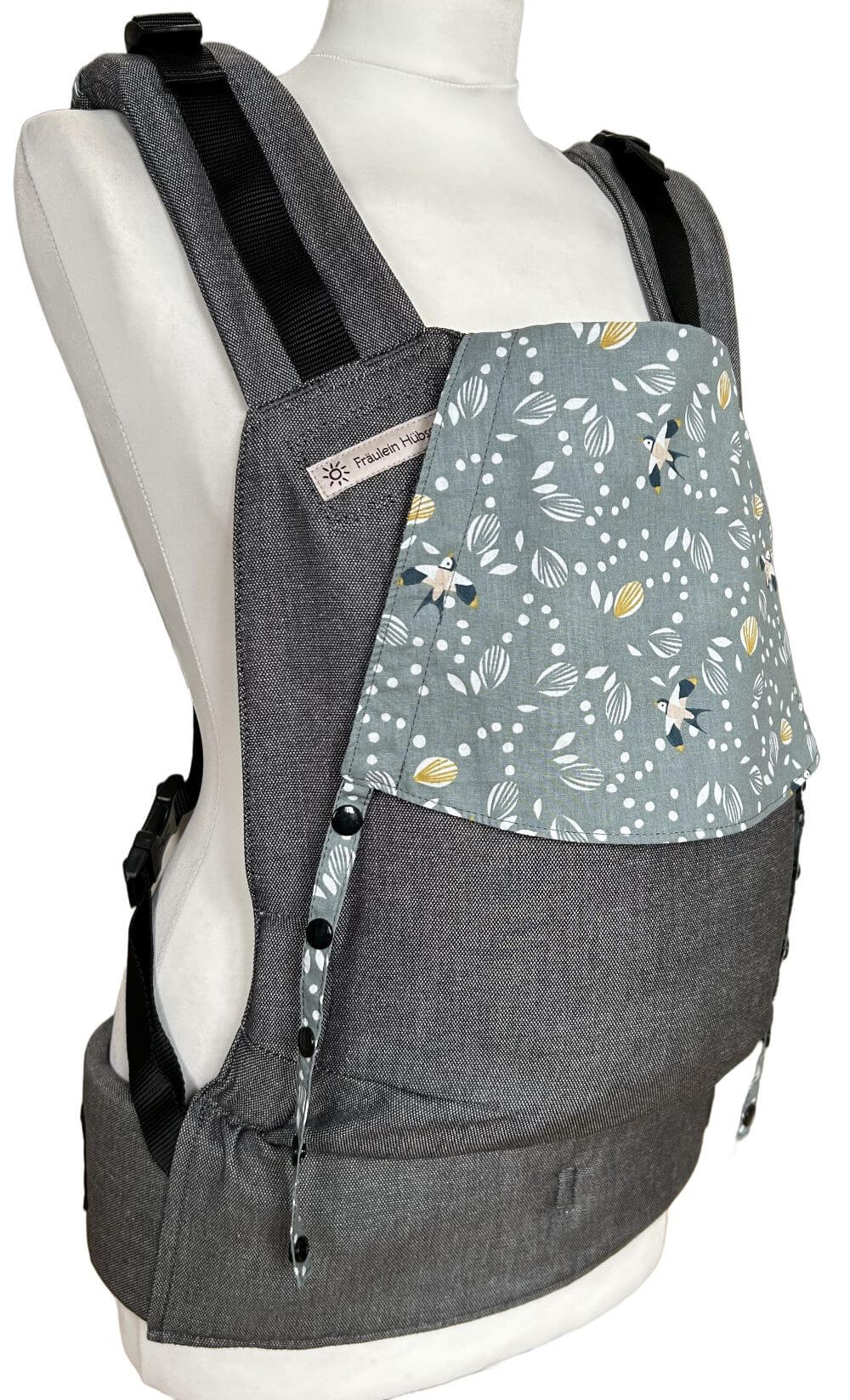 Fullbuckle Newborn Swallows Seitlich Fullbuckle Rucksacktrage Größe Newborn in Dunkelgrau mit einer Kopfstütze mit Schwalben auf Salbeigrün auf einer Schneiderbüste. Alle Gurte schließen mit Schnallen wie bei einem Rucksack, kein Binden.