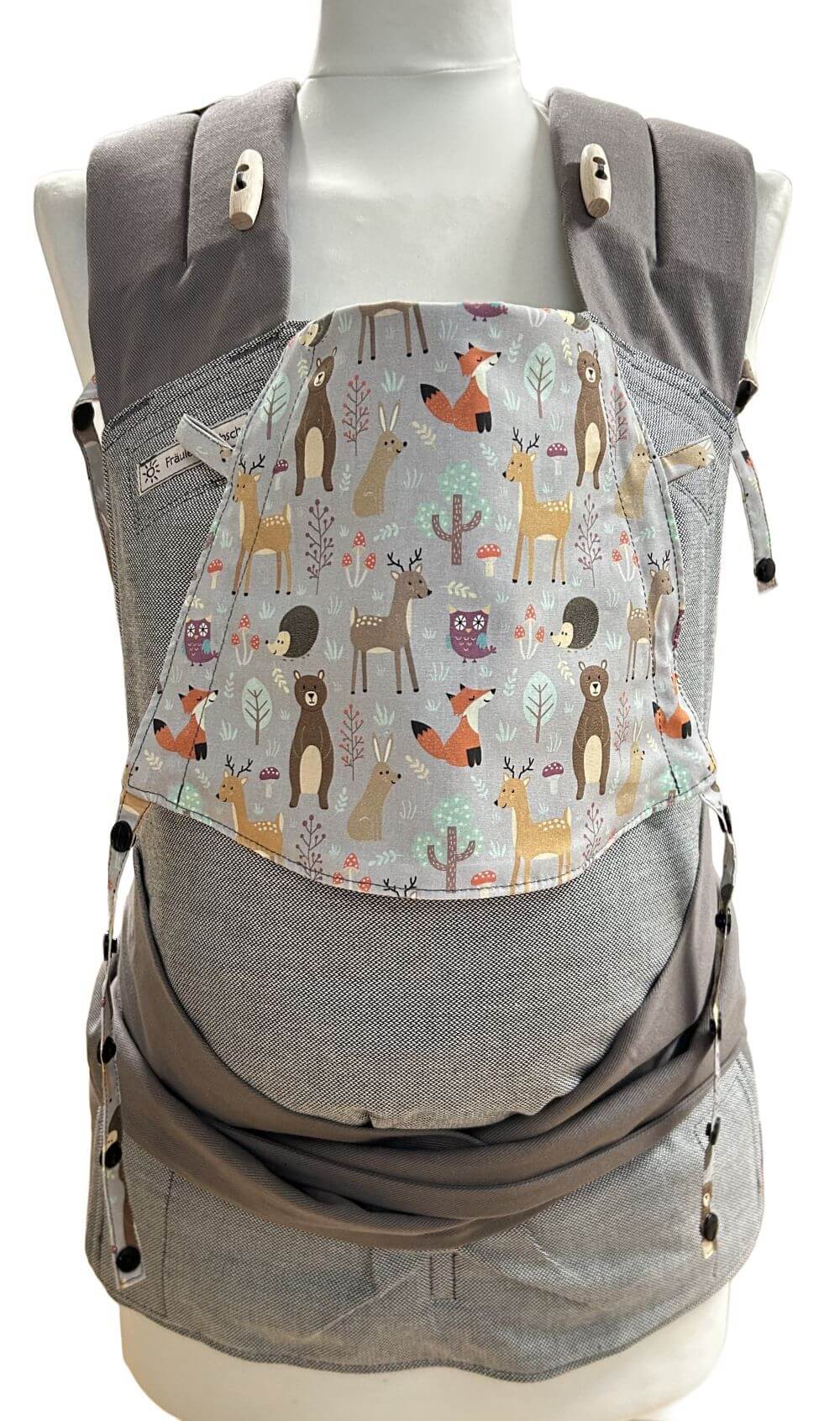 Mei Tai Babysize Zauberwald frontal Fräulein Hübsch Babytrage Mei Tai Babysize in Hellgrau mit bunter Kopfstütze im Waldtier-Design auf hellgrauem Hintergrund, präsentiert auf einer Schneiderpuppe.