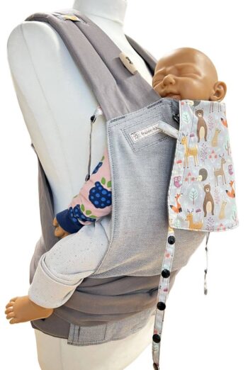 Fräulein Hübsch Babytrage Mei Tai Babysize in Hellgrau mit bunter Kopfstütze im Waldtier-Design auf hellgrauem Hintergrund, präsentiert auf einer Schneiderpuppe mit Demopuppe zur Veranschaulichung der Trageweise.