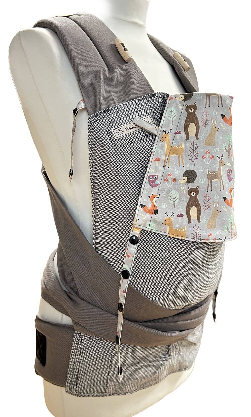 Mei Tai Babysize Zauberwald seitlich Fräulein Hübsch Babytrage Mei Tai Babysize in Hellgrau mit bunter Kopfstütze im Waldtier-Design auf hellgrauem Hintergrund, präsentiert auf einer Schneiderpuppe.