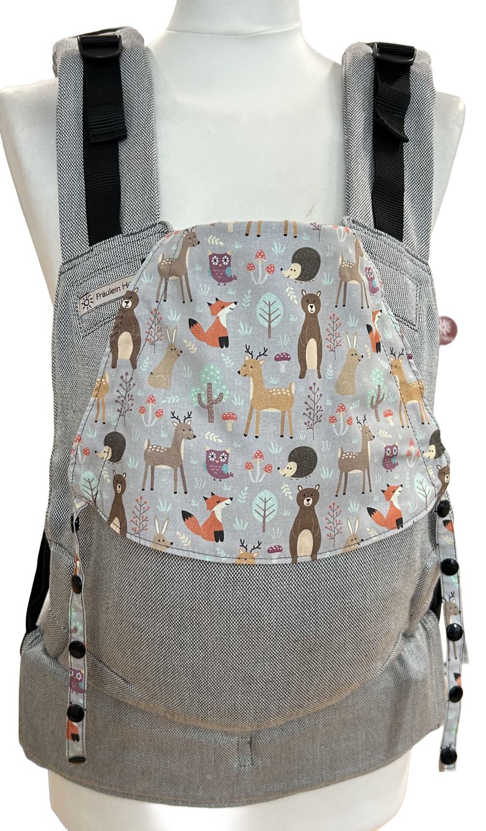 Fräulein Hübsch Fullbuckle Newborn in Hellgrau mit bunter Kopfstütze, auf der bunte Waldtiere auf hellgrauem Grund abgebildet sind. Die Trage wird auf einer Schneiderpuppe gezeigt.